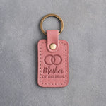 Parisian Blossom pink leather keychain debossed with two interlocked wedding rings above Mother and OF THE BRIDE, on gray concrete