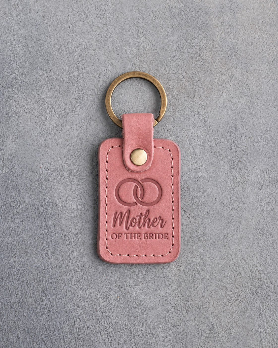 Parisian Blossom pink leather keychain debossed with two interlocked wedding rings above Mother and OF THE BRIDE, on gray concrete