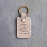 Gray sand leather keychain debossed with two interlocked wedding rings above Mother and OF THE GROOM, on gray concrete