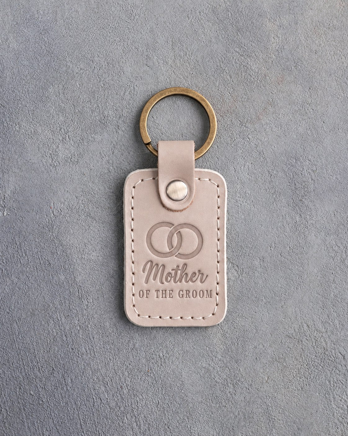 Gray sand leather keychain debossed with two interlocked wedding rings above Mother and OF THE GROOM, on gray concrete