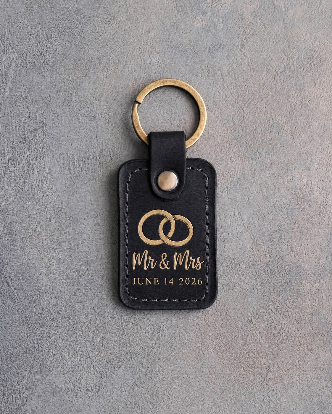 Black leather keychain stamped in gold foil with two interlocked wedding rings above Mr & Mrs and JUNE 14 2026, on gray concrete