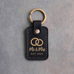 Black leather keychain stamped in gold foil with two interlocked wedding rings above Mr & Mrs and EST 2026, on gray concrete