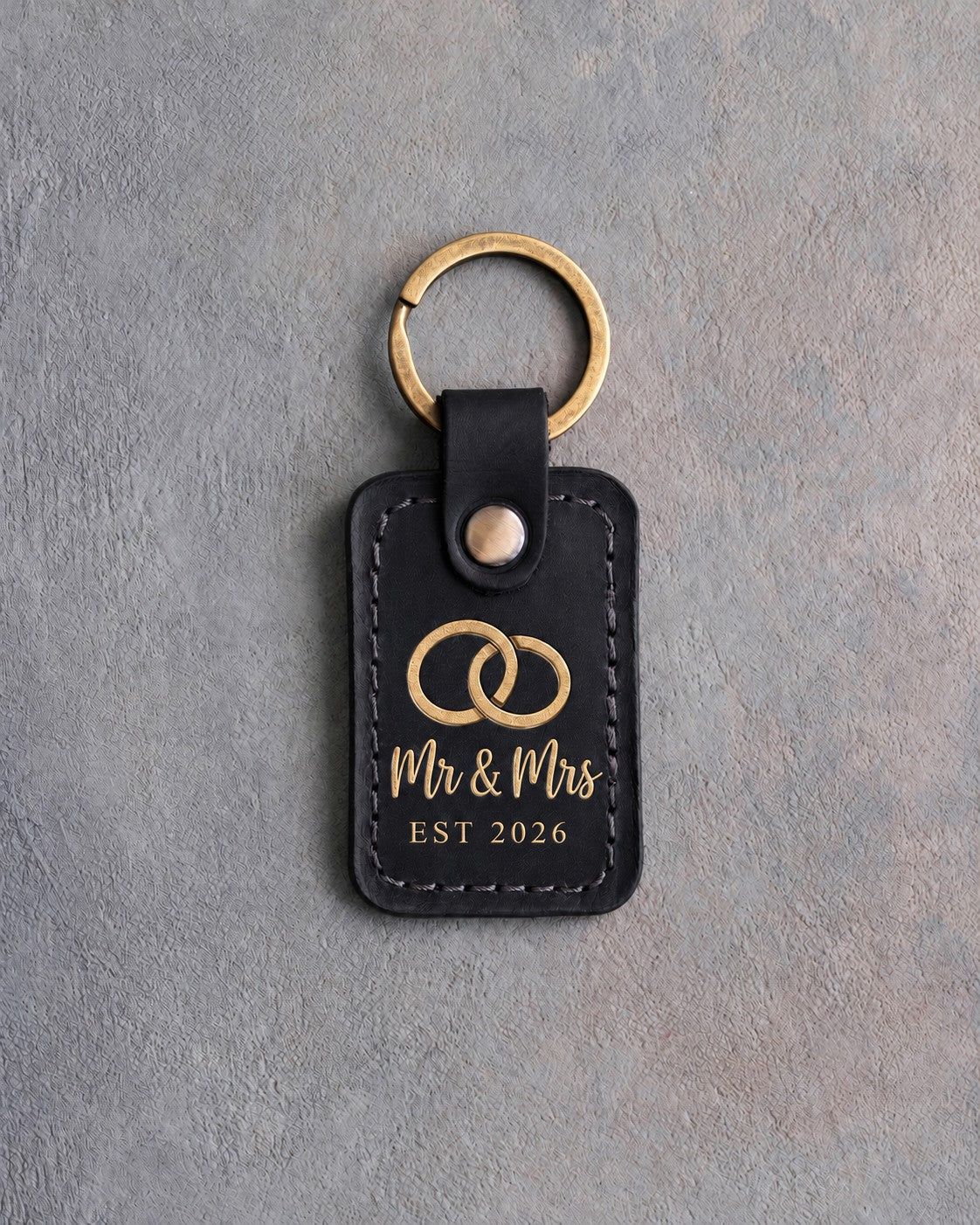 Black leather keychain stamped in gold foil with two interlocked wedding rings above Mr & Mrs and EST 2026, on gray concrete