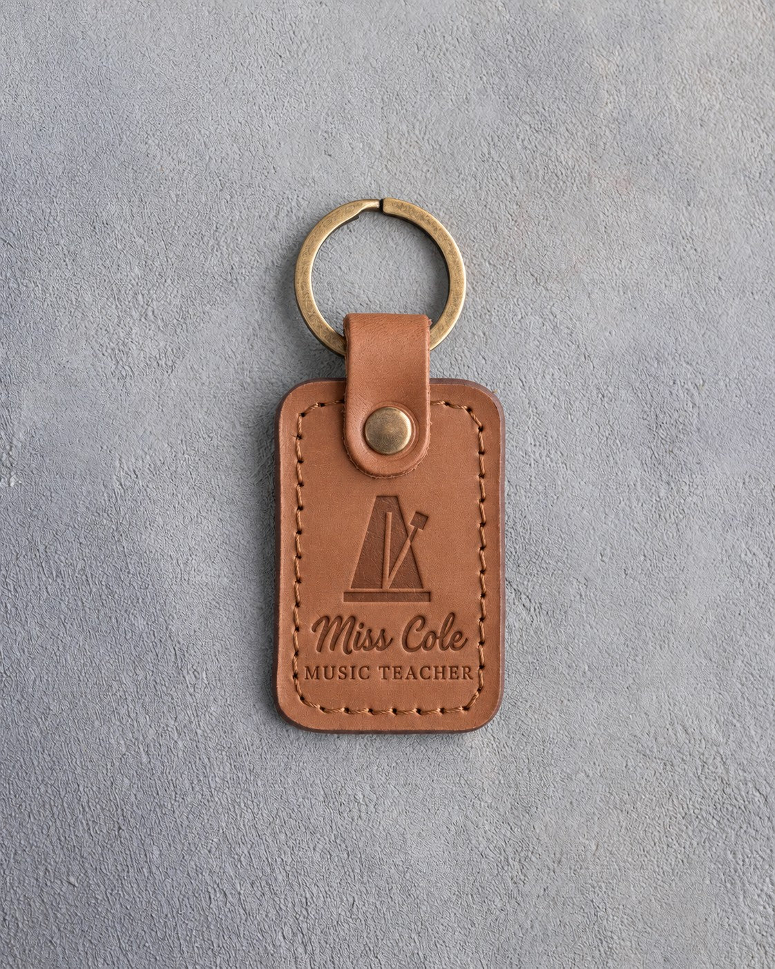 Caramel leather keychain debossed with a pyramid metronome above the name Miss Cole and MUSIC TEACHER, on gray plaster