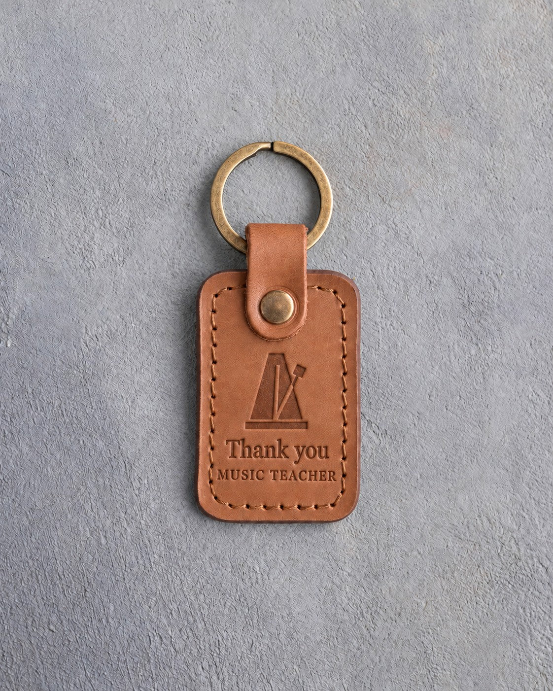 Caramel leather keychain debossed with a pyramid metronome above the words Thank you and MUSIC TEACHER, on gray concrete