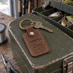 Sicilian Brown Lars MUSKY keychain in full-grain leather on the lid of an olive-green steel tackle box in a garage, lures in the trays behind