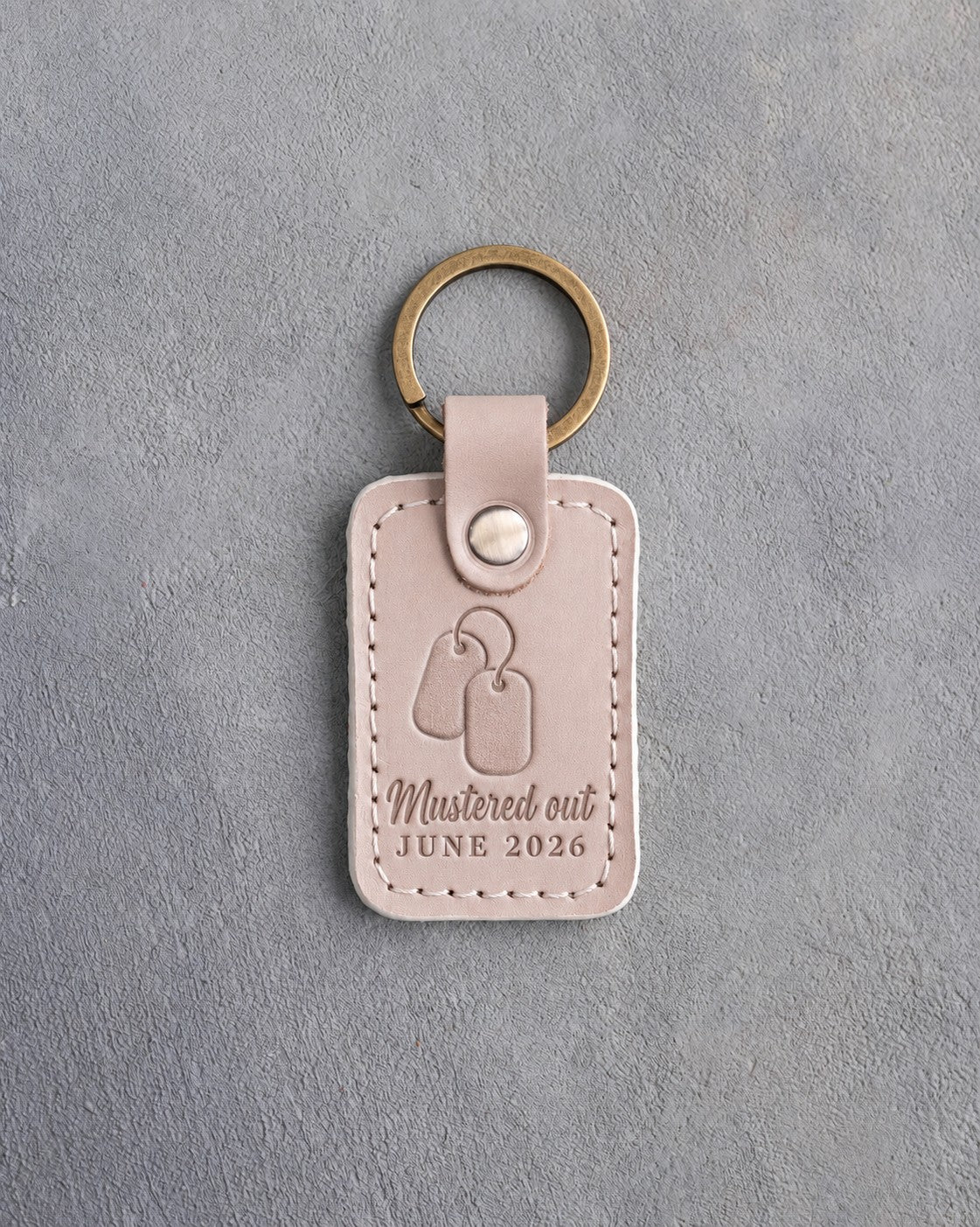 Gray sand leather keychain debossed with a pair of dog tags above the words Mustered out and JUNE 2026, on gray concrete