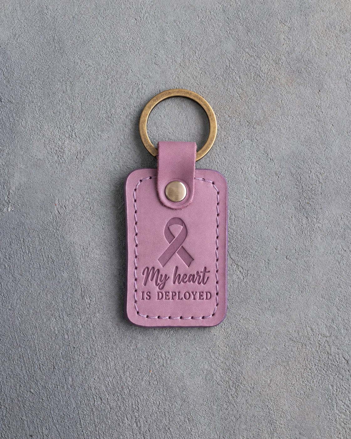 Lilac leather keychain debossed with a looped ribbon above the words My heart IS DEPLOYED, on gray concrete
