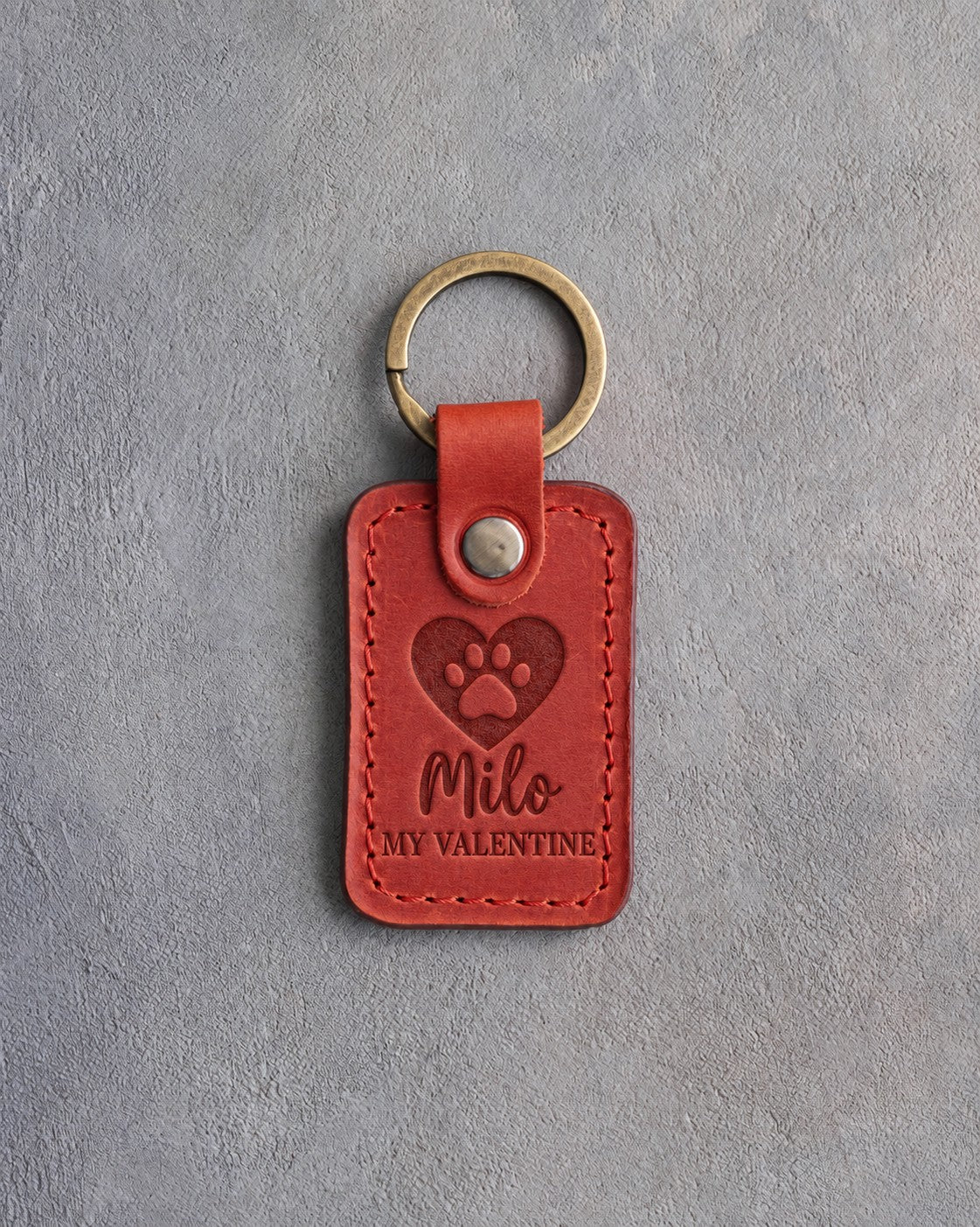 Poppy red leather keychain debossed with a heart holding a paw print above Milo and MY VALENTINE, on gray concrete