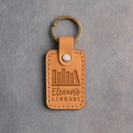 Indian Summer tan leather keychain debossed with a row of books on a shelf above Eleanor's and LIBRARY, on gray concrete