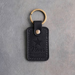 Black leather keychain debossed with a five-point star above the words Never FORGOTTEN, on gray concrete