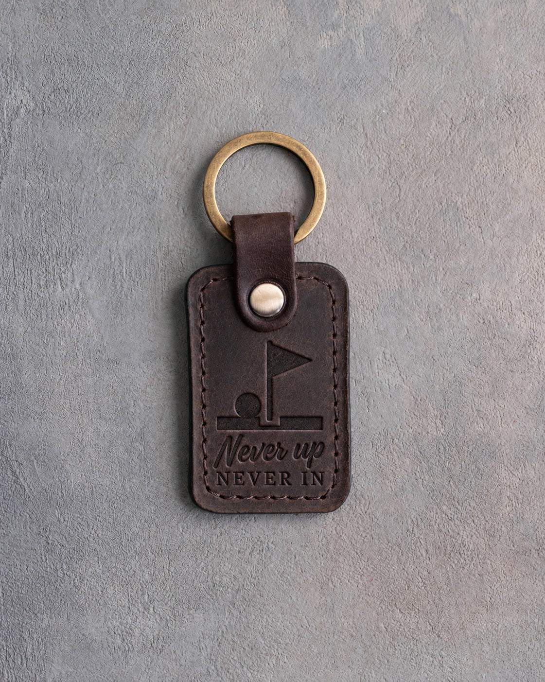 Dark espresso leather keychain debossed with a golf ball on the lip of the cup above Never up NEVER IN, on gray plaster