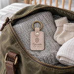 Pale gray New mom EST 2026 keychain clipped to a diaper bag strap on a nursery chair