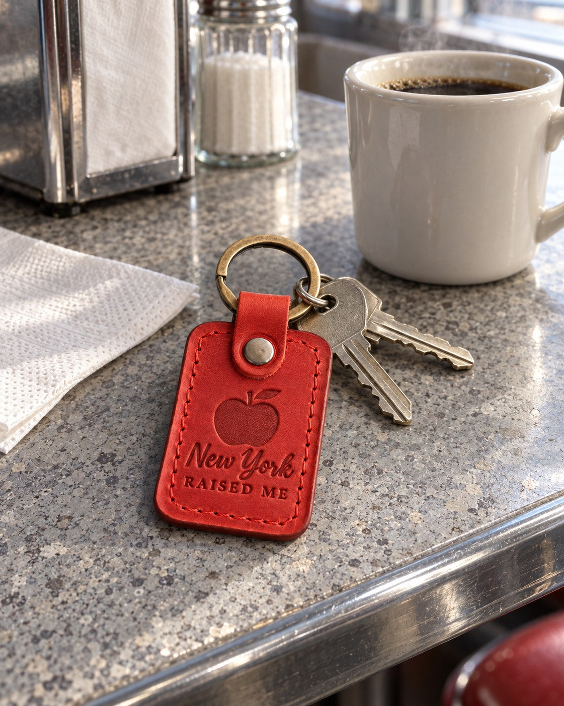 Red New York Raised Me keychain on apartment keys held up at a brownstone stoop on a bright afternoon