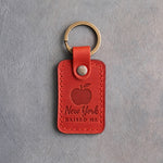 Poppy red leather keychain debossed with a solid apple above New York and RAISED ME, on gray concrete