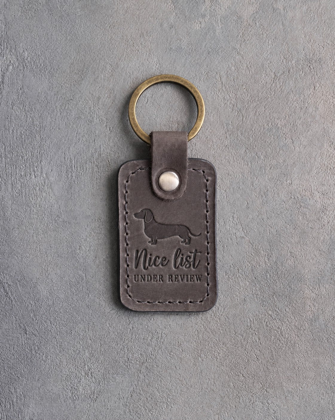 Country gray leather keychain debossed with a dachshund silhouette and the words Nice list UNDER REVIEW, on gray concrete