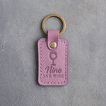 Lilac leather keychain debossed with a wine glass holding a golf ball above the words Nine AND WINE, on gray plaster