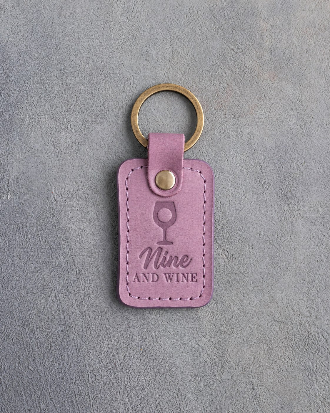 Lilac leather keychain debossed with a wine glass holding a golf ball above the words Nine AND WINE, on gray plaster