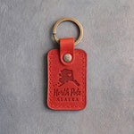 Poppy red leather keychain debossed with an Alaska silhouette and a heart at North Pole above North Pole and ALASKA, on gray concrete