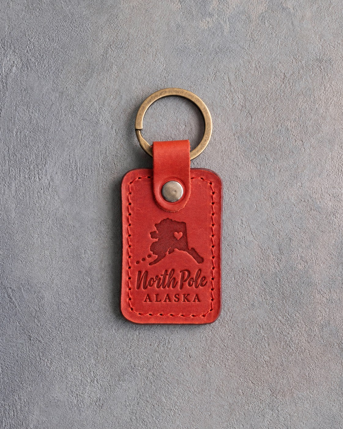 Poppy red leather keychain debossed with an Alaska silhouette and a heart at North Pole above North Pole and ALASKA, on gray concrete