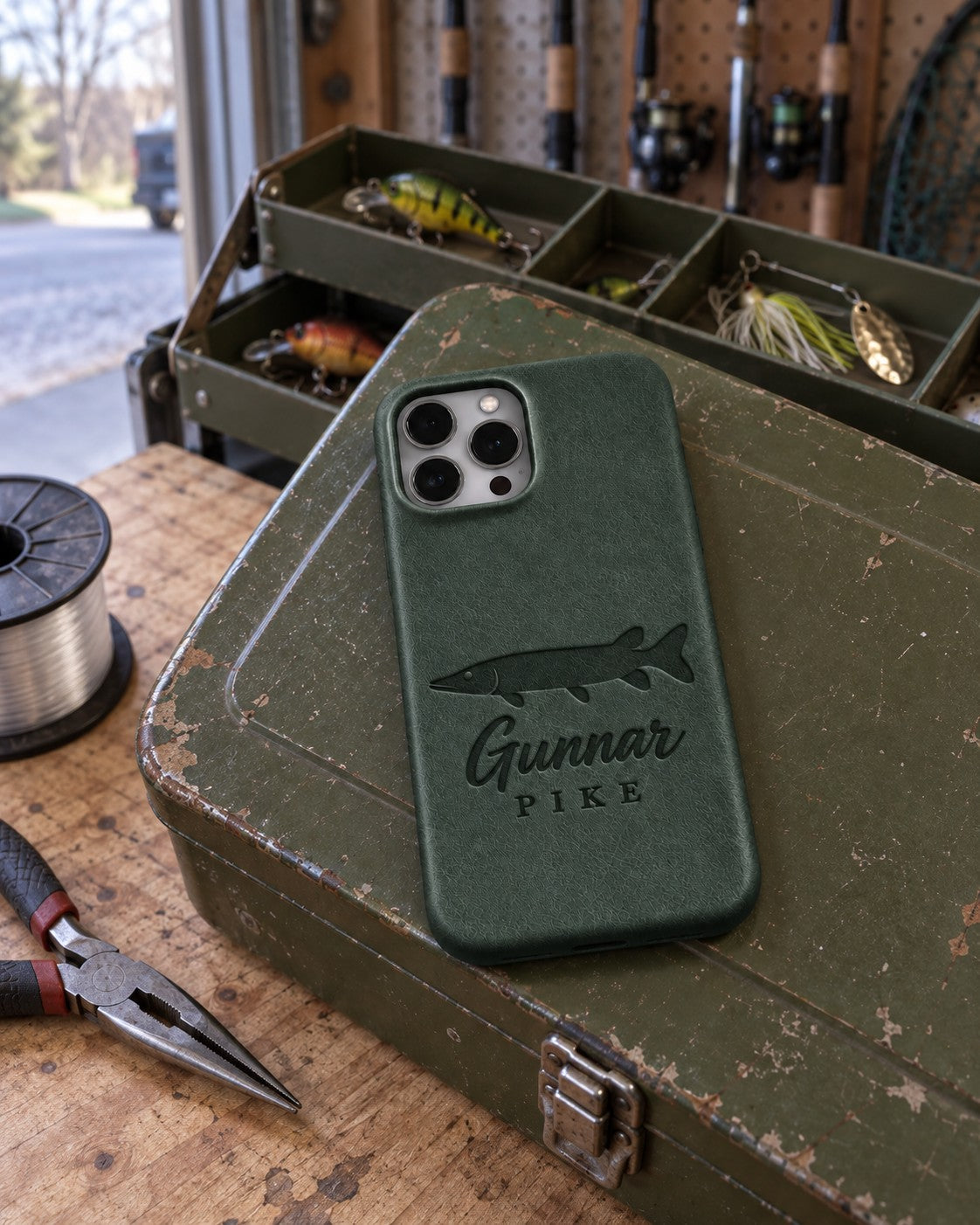 Forest Green Gunnar PIKE iPhone case in full-grain leather on the lid of an olive-green steel tackle box in a garage, lures in the trays behind