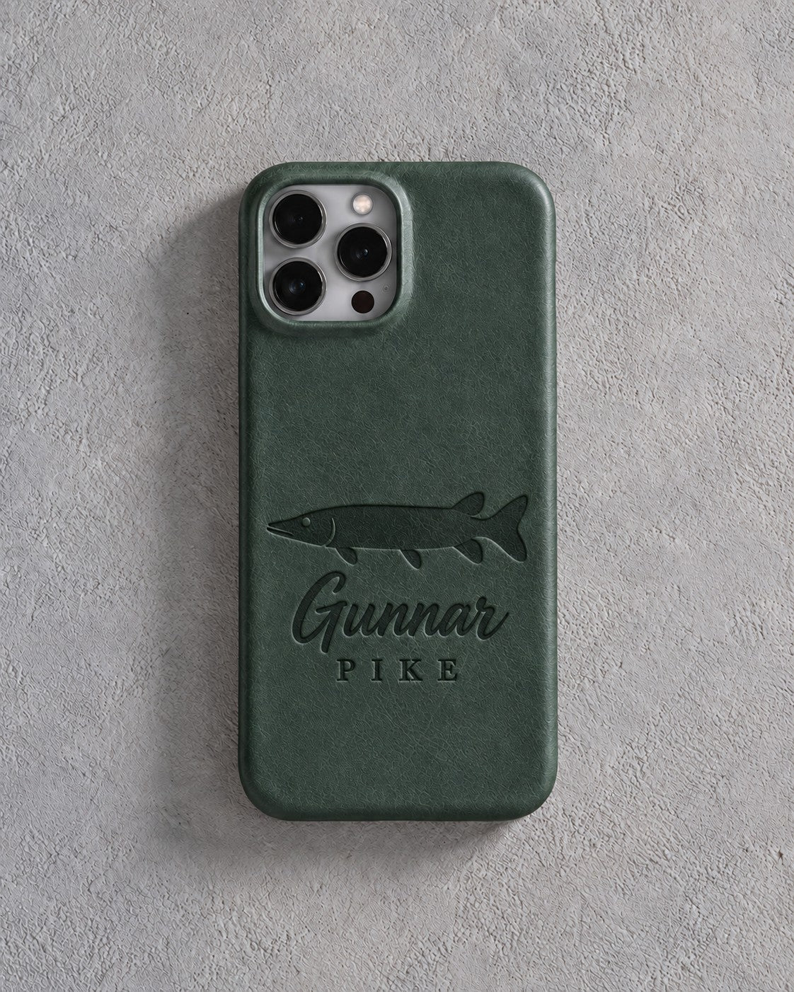 Back of a forest green leather iPhone case debossed with a northern pike above Gunnar and PIKE below the camera, on gray plaster