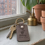 Gray Nothing Grows Here keychain on a windowsill beside a small empty terracotta pot