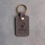 Country gray leather keychain debossed with a pot holding one drooping leaf above Nothing GROWS HERE, on gray concrete