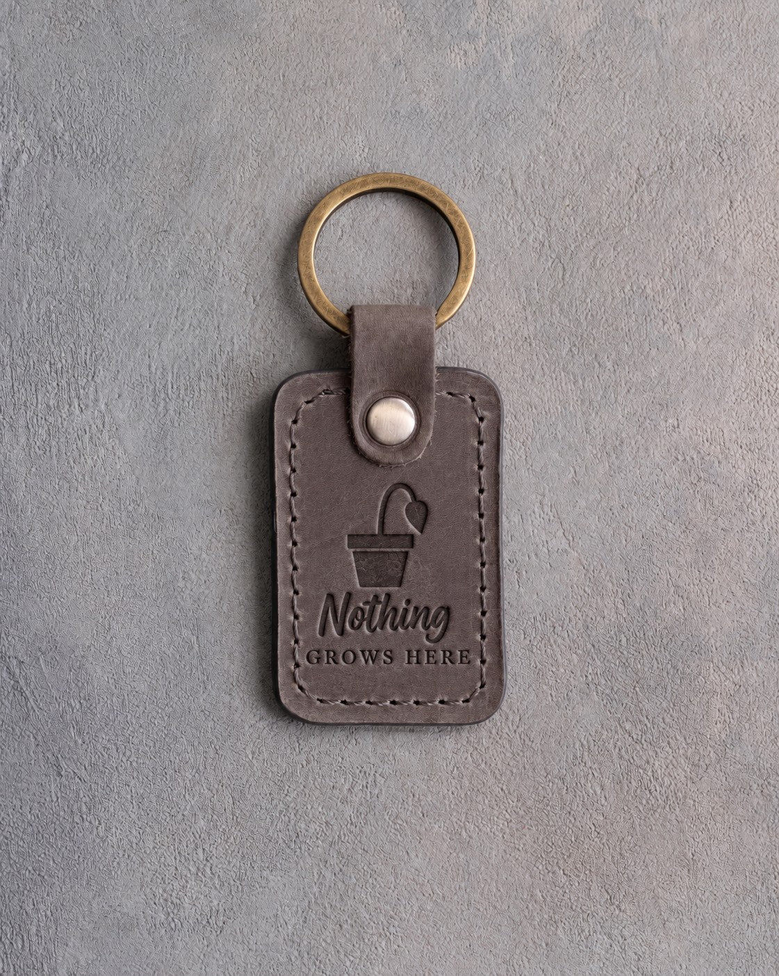 Country gray leather keychain debossed with a pot holding one drooping leaf above Nothing GROWS HERE, on gray concrete