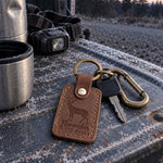 November Is for Bucks keychain in brown leather lying on frosted oak leaves beside a grunt call