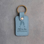 Turquoise leather keychain debossed with two toasting champagne flutes above the words Off to the HONEYMOON, on gray concrete