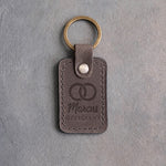 Country gray leather keychain debossed with two interlocked wedding rings above the name Marcus and OFFICIANT, on gray concrete