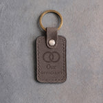 Country gray leather keychain debossed with two interlocked wedding rings above Our and OFFICIANT, on gray concrete