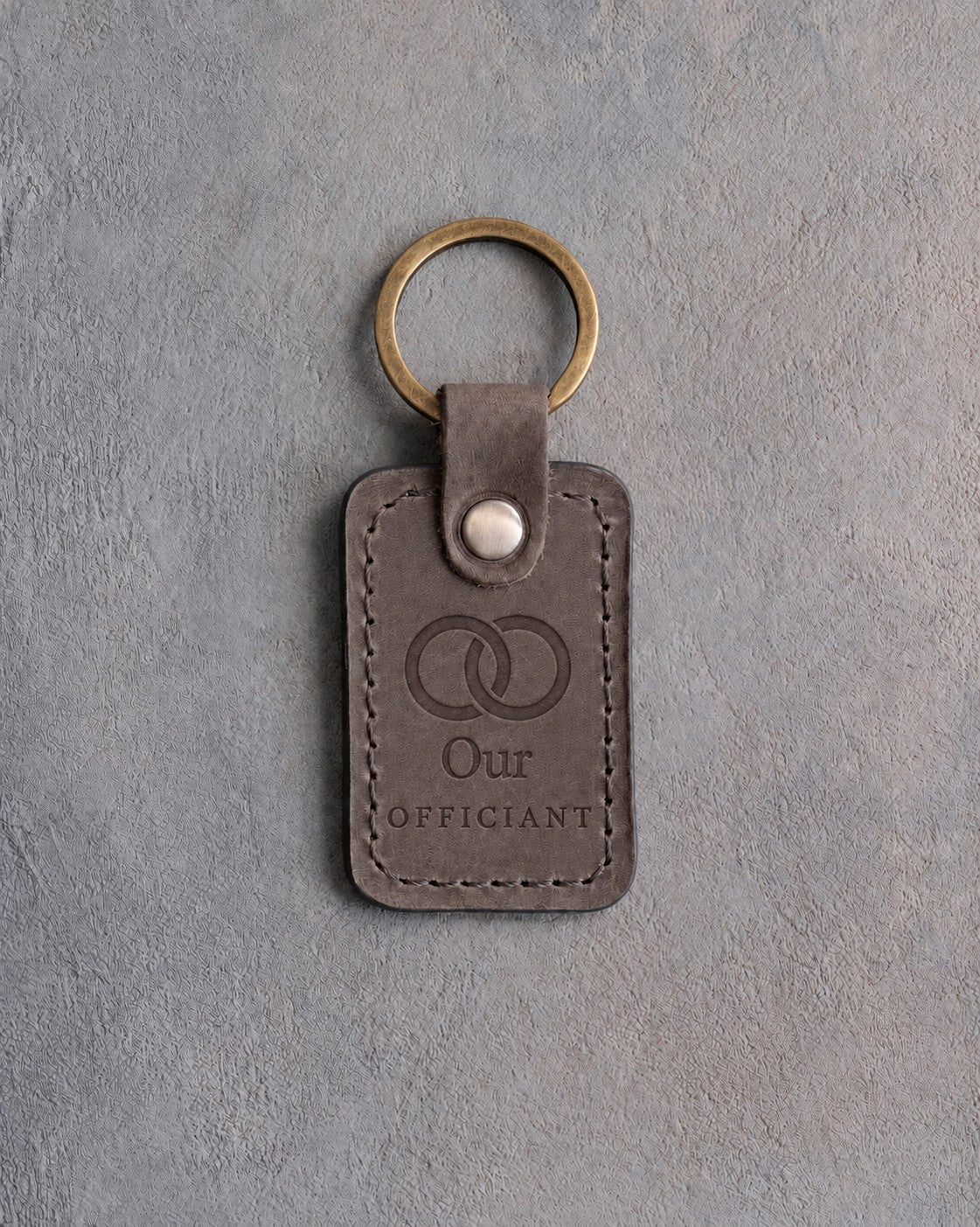 Country gray leather keychain debossed with two interlocked wedding rings above Our and OFFICIANT, on gray concrete