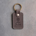 Country gray leather keychain debossed with an erupting geyser above On time and YELLOWSTONE, on gray concrete