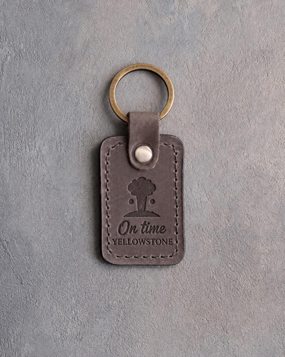 Country gray leather keychain debossed with an erupting geyser above On time and YELLOWSTONE, on gray concrete
