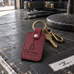 One More Guitar keychain in wine red leather on a house key hanging beside a wall of hung guitars