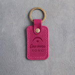 Fuchsia pink leather keychain debossed with a fermata above the words One more SONG, on gray plaster