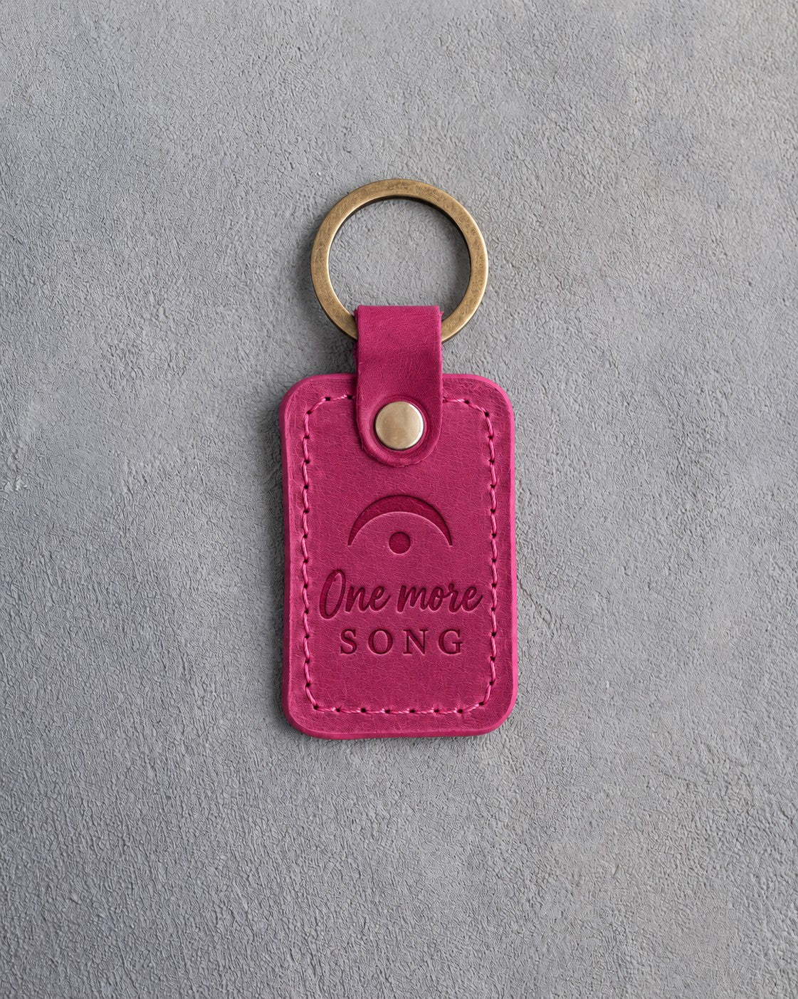 Fuchsia pink leather keychain debossed with a fermata above the words One more SONG, on gray plaster