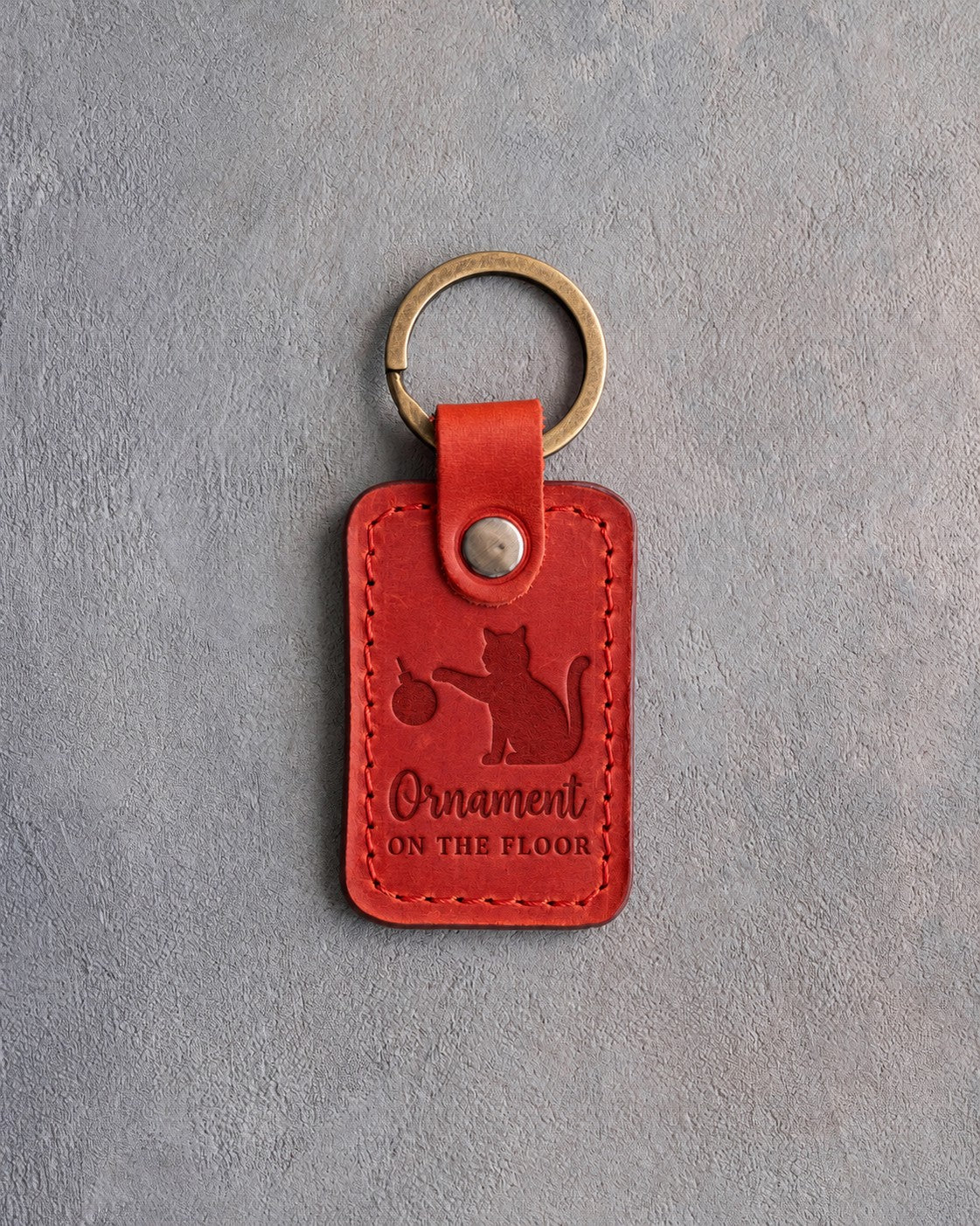 Poppy red leather keychain debossed with a cat knocking a Christmas bauble off its paw above the words Ornament ON THE FLOOR