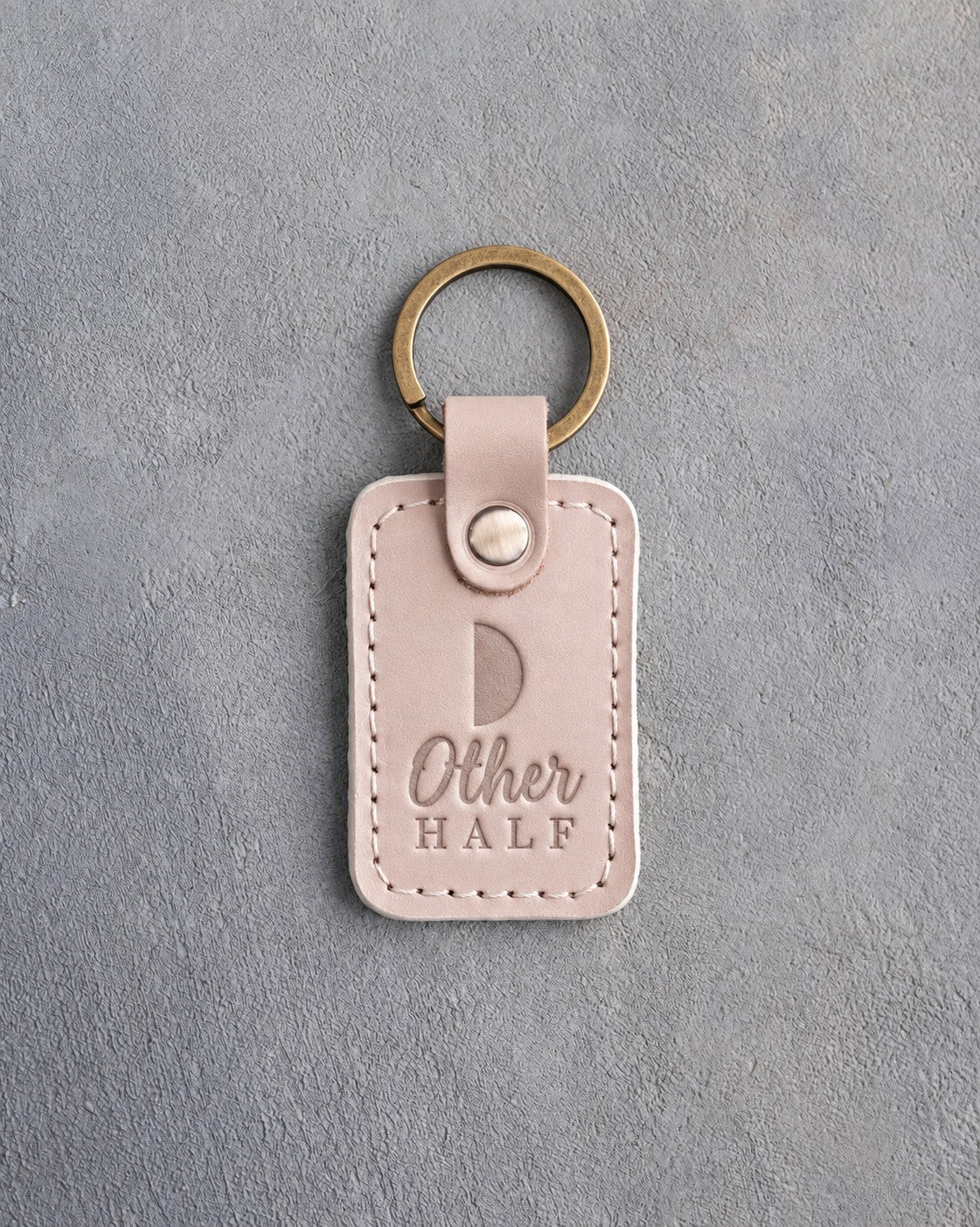 Gray sand leather keychain debossed with a first quarter half moon above the words Other HALF, on gray concrete