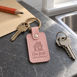 Pink Our First Christmas keychain on a new house key resting under a small Christmas tree in an empty living room