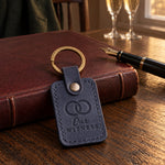 Our WITNESS keychain in navy leather on a dark oak table before a closed leather register and a fountain pen