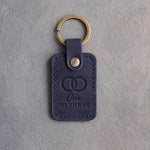 Blueberry navy leather keychain debossed with two interlocked wedding rings above the words Our WITNESS, on gray concrete