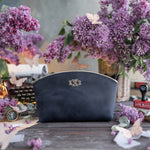 Navy blueberry leather makeup pouch with an ornate gold monogram, lilacs behind