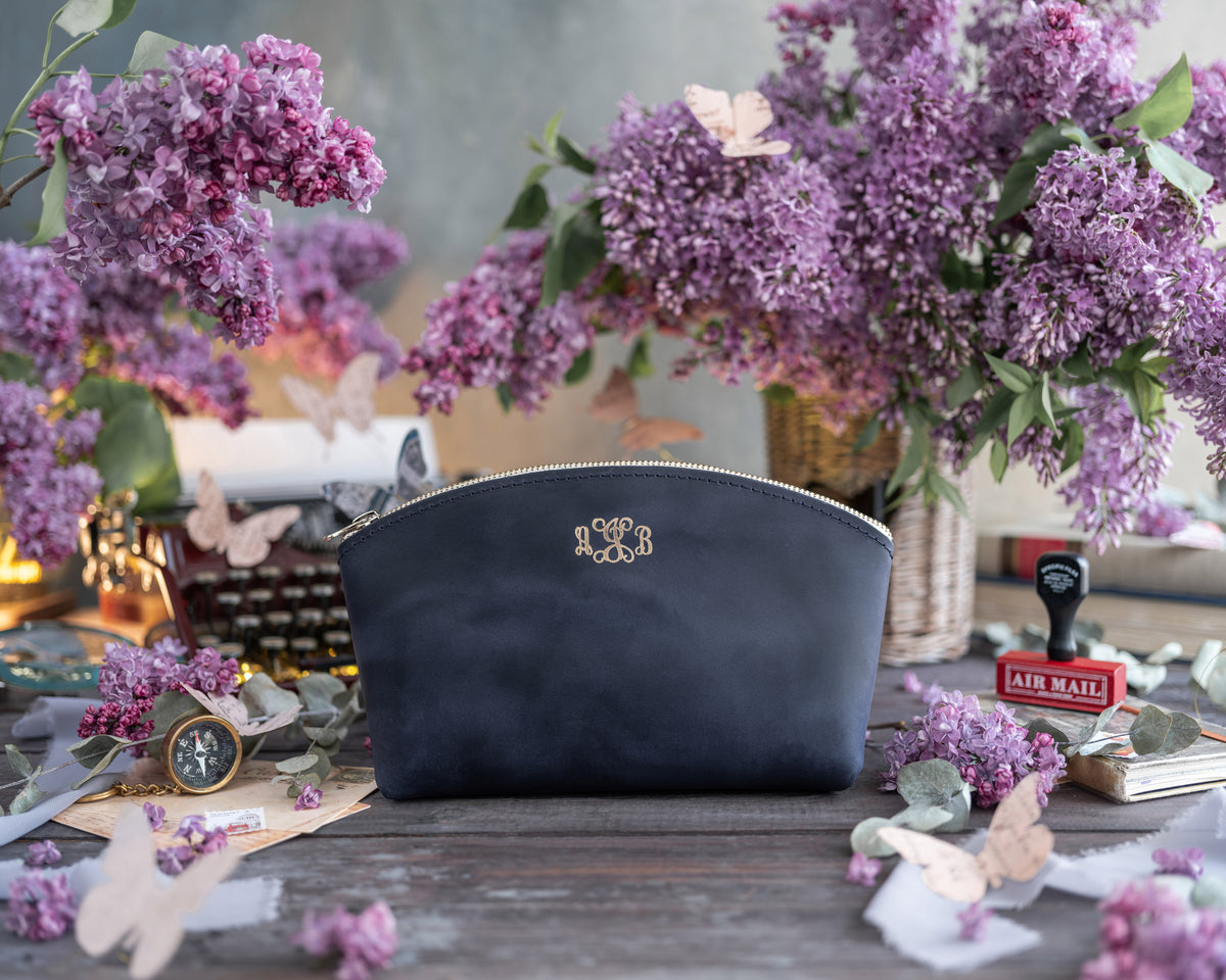 Navy blueberry leather makeup pouch with an ornate gold monogram, lilacs behind