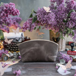 Gray leather makeup bag with a small gold monogram on a dark rustic table with lilacs