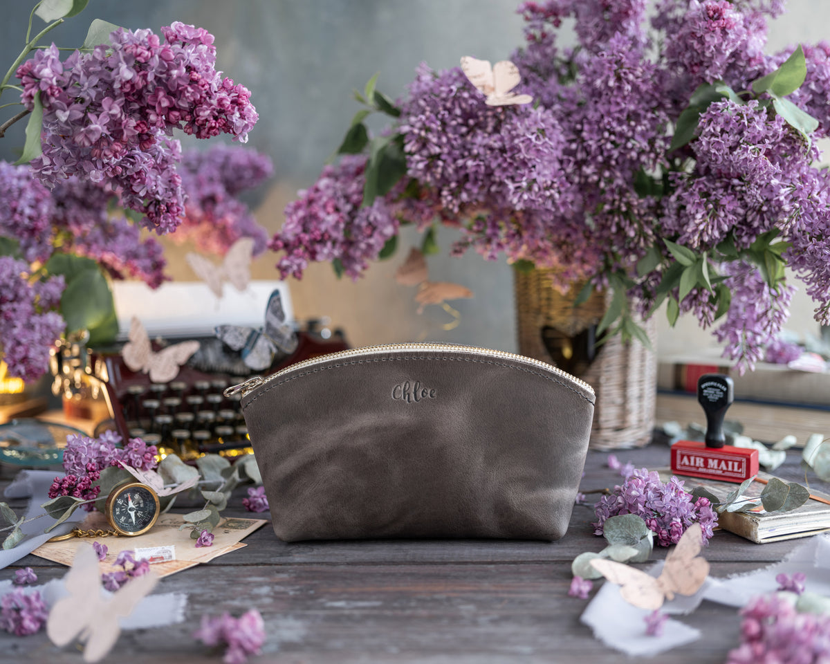 Gray leather makeup bag with a small gold monogram on a dark rustic table with lilacs