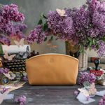 Golden tan leather makeup bag with a small gold monogram, lilacs on a rustic table
