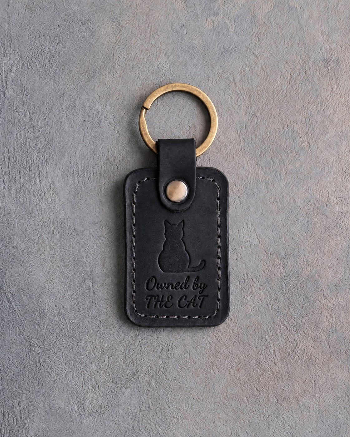Black leather keychain with a debossed cat seen from behind above the words Owned by and THE CAT, on gray concrete